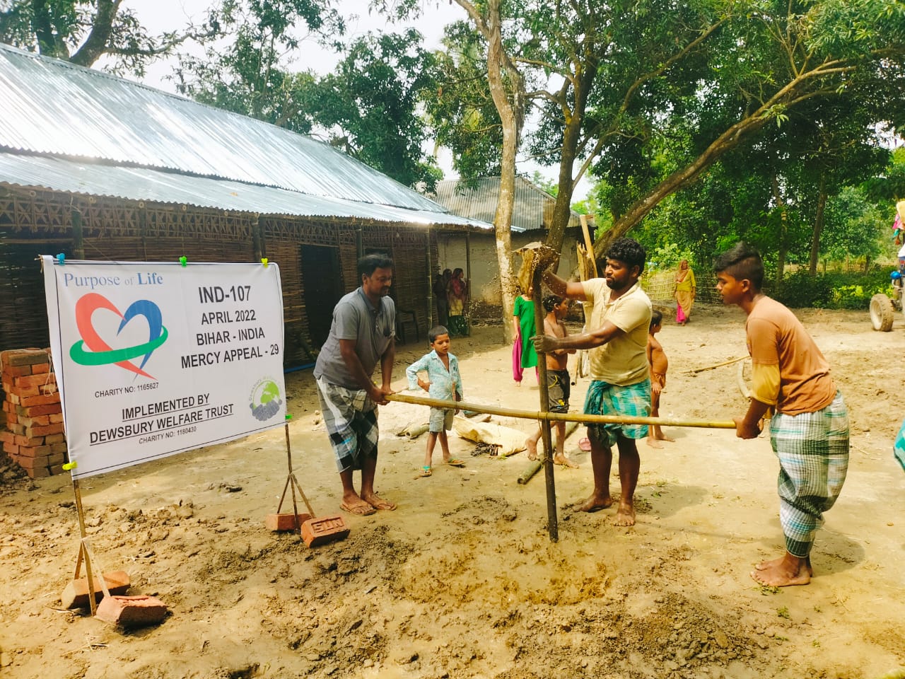 People working on constructing a water pump in India with a 'Purpose of Life' banner in the background.