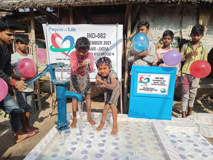 Children playing with balloons near a water pump and 'Purpose of Life' sign in a rural setting.