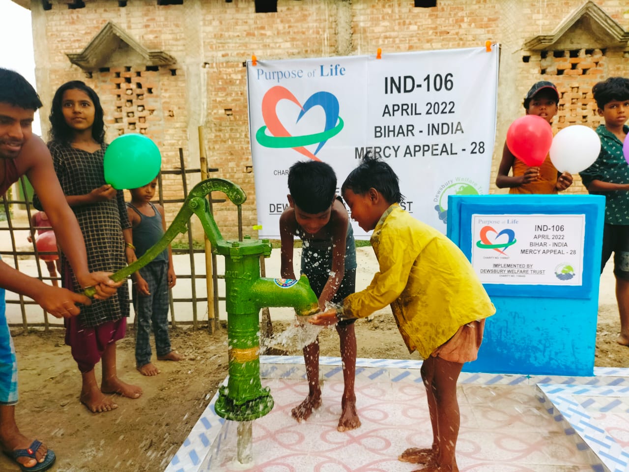 Children using a water pump with a 'Purpose of Life' banner in the background.