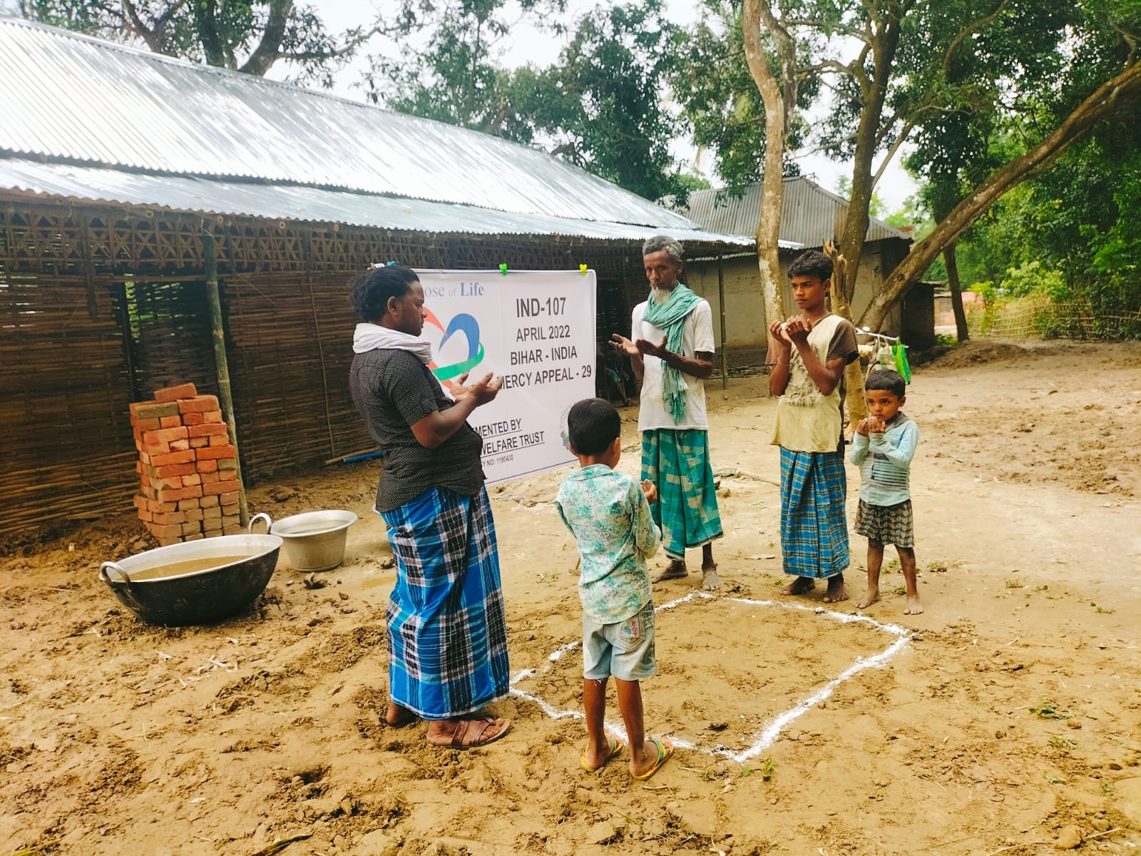 People standing outdoors with a banner and children making dua and prayer in a rural setting