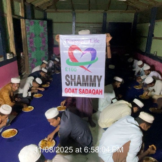 Group of people sitting on the floor in a room with a large sign promoting 'Shammy Goat Sadaqah'.